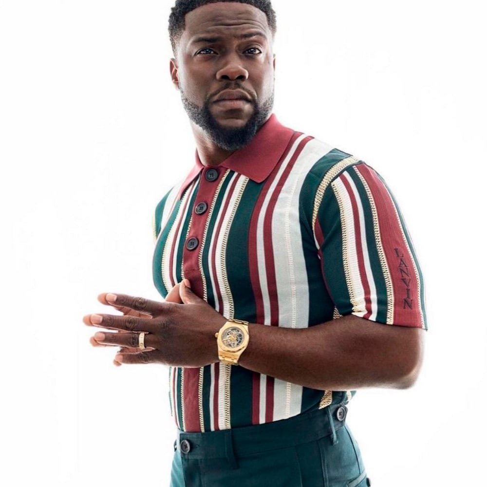 A man with short hair and a beard poses against a white background, wearing a striped short-sleeve shirt with red, green, and white vertical stripes, dark green pants, a gold watch, and a ring on his left hand.