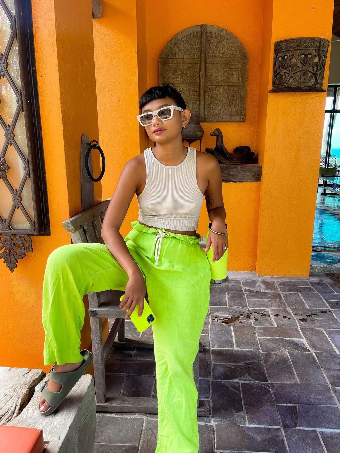 A woman in a beige sleeveless top and neon green pants sits on a wooden bench against a bright orange wall, holding a matching neon tumbler and phone, wearing sunglasses and sandals.