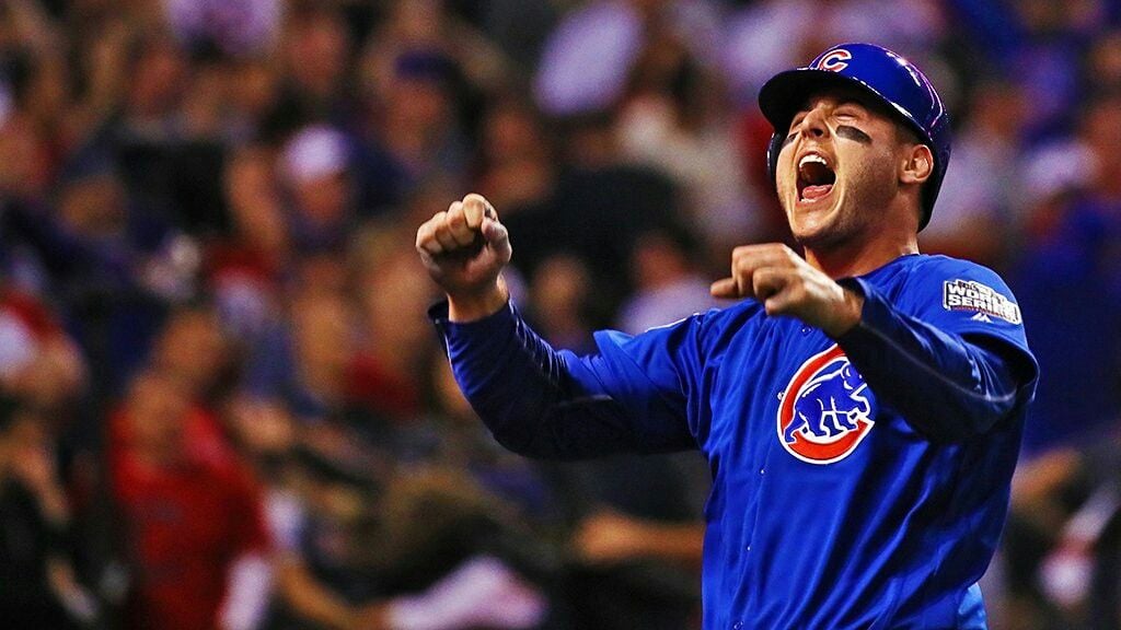 A Chicago Cubs baseball player in a blue uniform and helmet celebrates with arms raised and mouth open during a game, wearing black eye paint. The crowd is blurred in the background.