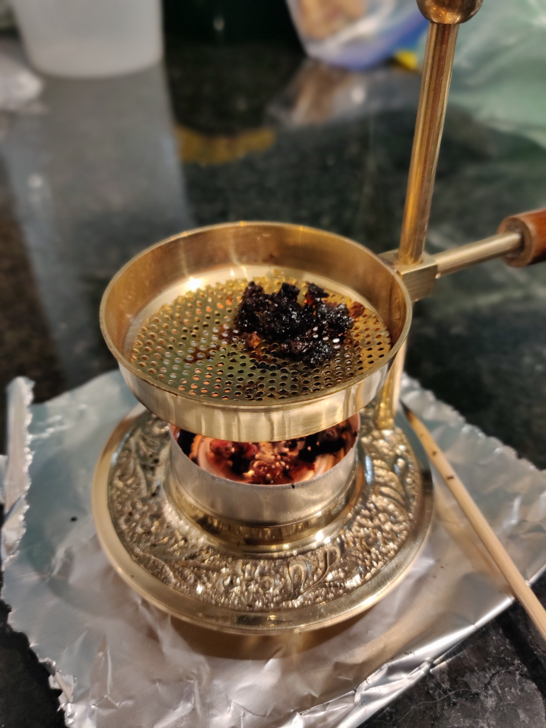 A brass incense burner with a perforated tray holds smoldering incense over hot coals. The burner rests on a foil-covered surface, with a metal handle and intricate engraved designs.