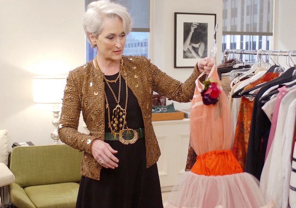 A stylish older woman with short white hair examines a peach and red dress in a modern office filled with racks of clothing. She wears a gold-sequined jacket, layered necklaces, and looks thoughtfully at the dress.