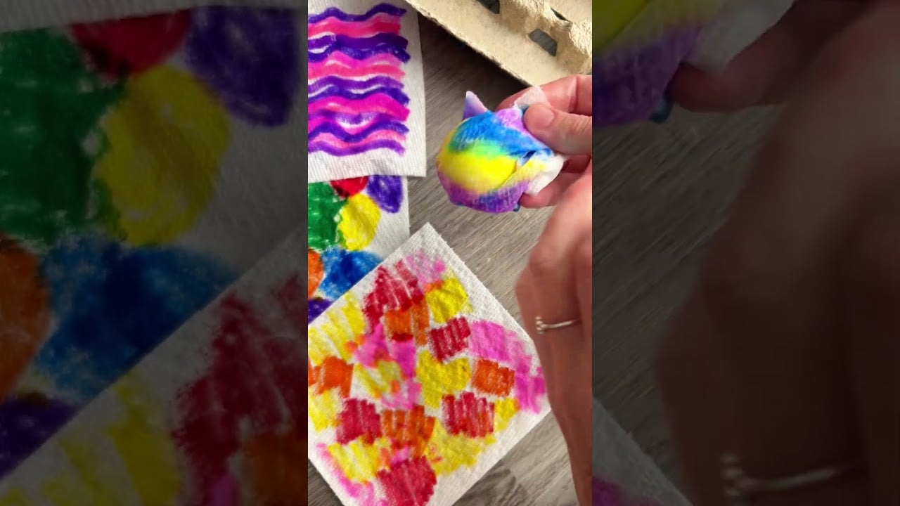 A person holds a small folded napkin colored with rainbow markers, surrounded by other vibrant, hand-decorated napkins on a wooden surface.