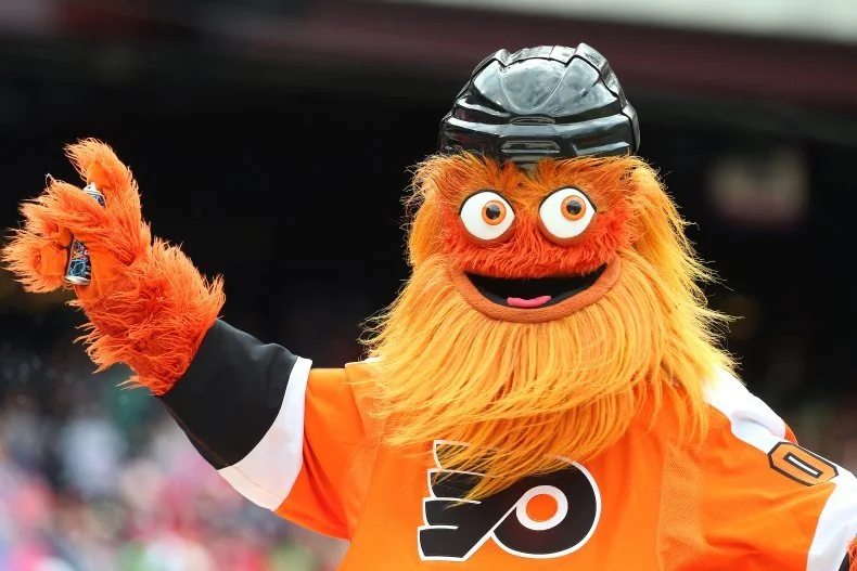A fluffy orange mascot with big googly eyes, a wide mouth, and a hockey helmet wears a Philadelphia Flyers jersey while waving.
