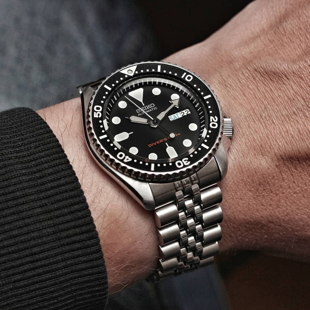 A Seiko automatic diver's watch with a black dial, round white markers, and a metal jubilee bracelet is worn on a person's left wrist with a dark sweater sleeve visible. The day and date are displayed at 3 o'clock.