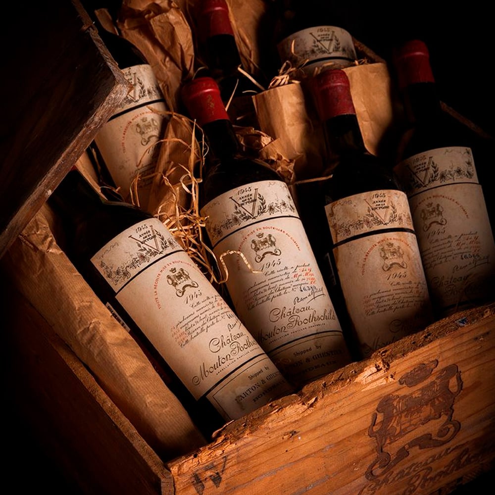 A wooden crate filled with several vintage bottles of Château Mouton Rothschild wine, some wrapped in brown paper and straw for protection, with old labels and red foil tops visible.