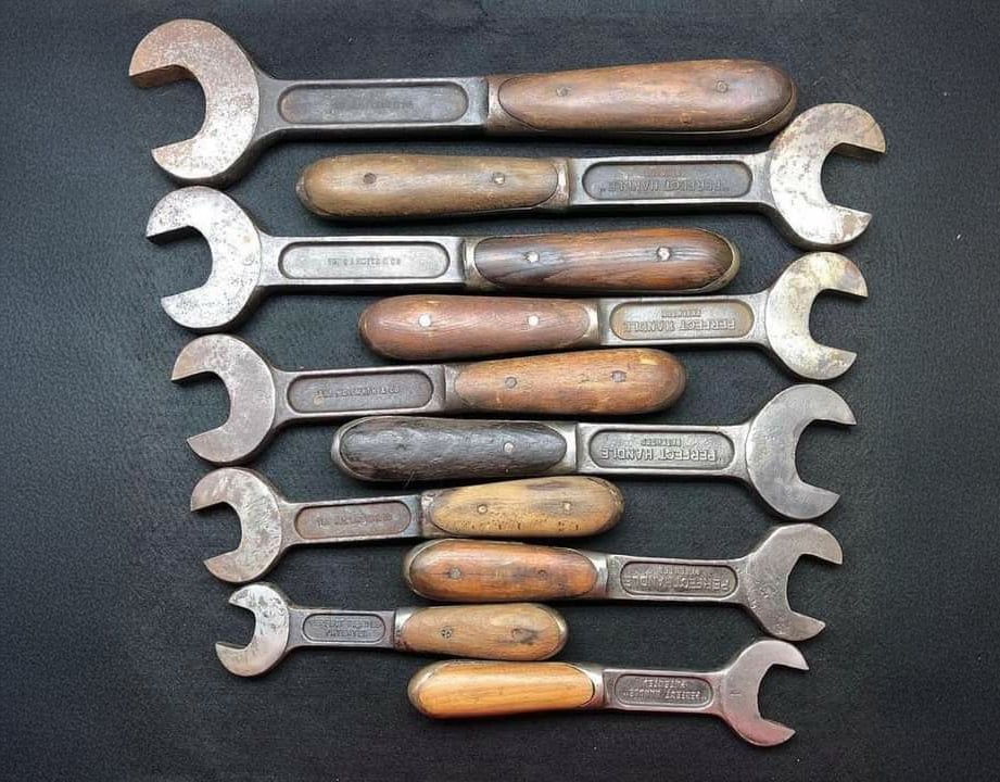 Nine vintage wrenches with wooden handles are arranged horizontally on a dark surface, forming a neat pattern. The metal heads of the wrenches face alternating directions.