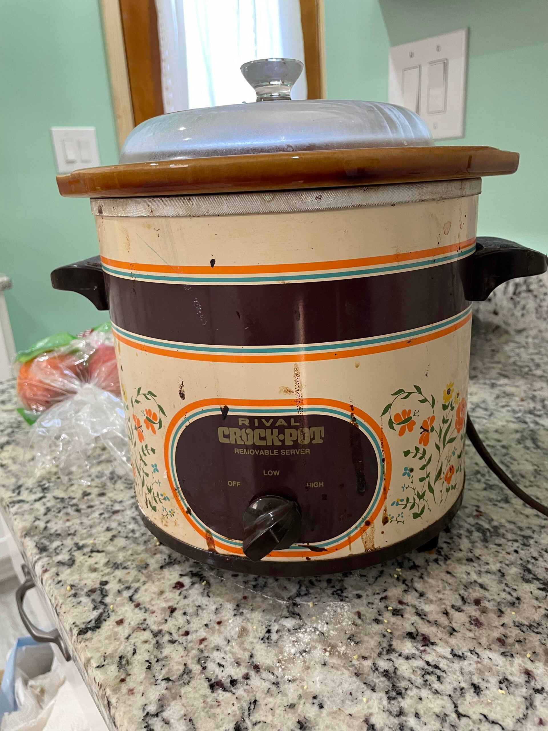 A vintage Rival Crock-Pot slow cooker with floral designs sits on a granite countertop. The Crock-Pot has a brown and beige body, a knob with off, low, and high settings, and a ceramic lid.
