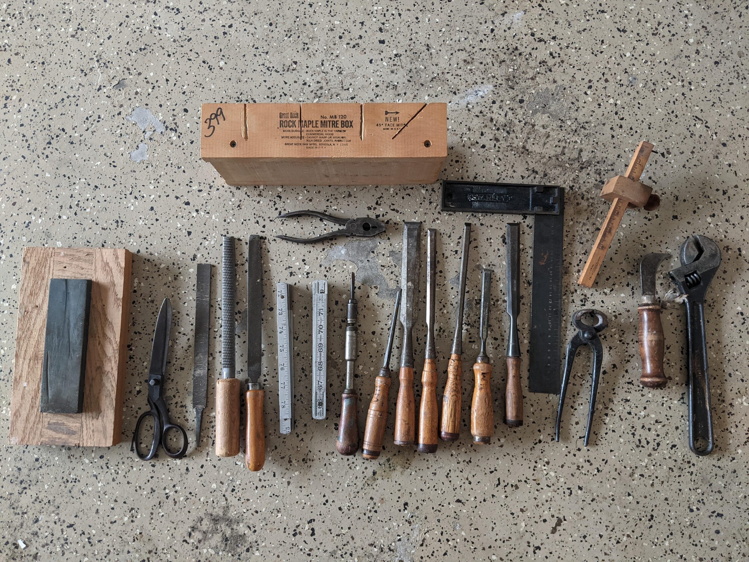 A variety of woodworking hand tools including chisels, files, rulers, pliers, scissors, a saw, a mallet, and a wooden box are neatly arranged on a speckled surface.