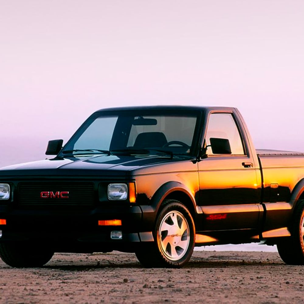 A black GMC pickup truck parked on sandy ground at sunset, with orange sunlight reflecting off its body and a clear sky in the background.
