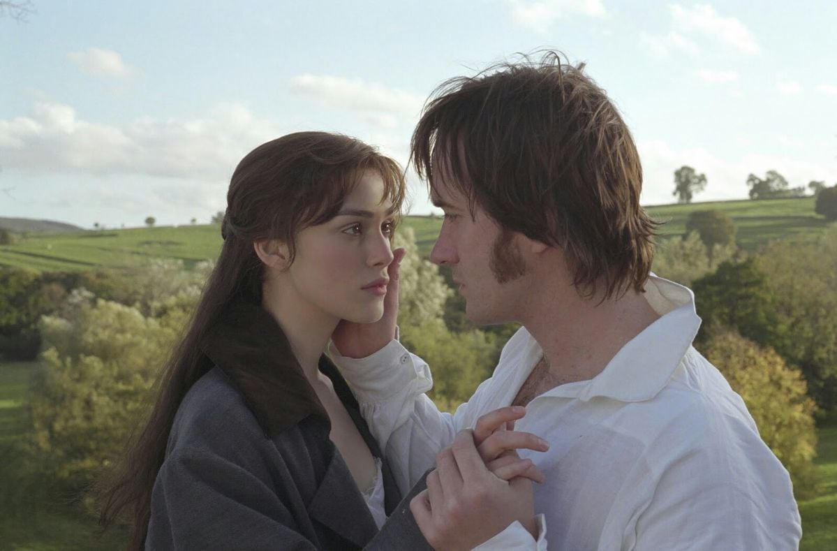 A man and woman stand closely together outdoors, gazing into each other’s eyes. He gently holds her face and hand. Both have serious, emotional expressions, with a green, sunlit countryside landscape in the background.
