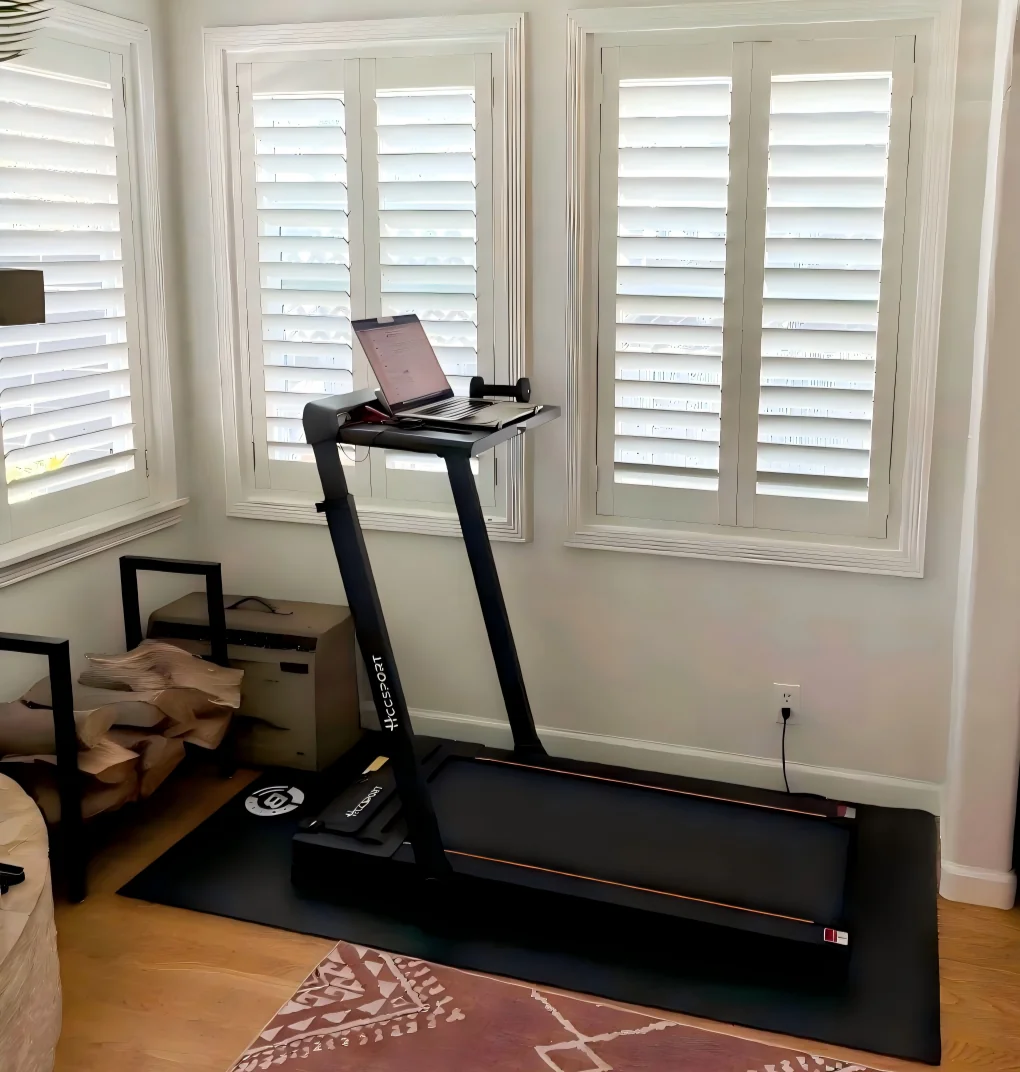 A treadmill with a laptop on its console is set up in a bright room with white shuttered windows, surrounded by a mat, some boxes, and a patterned rug on the wooden floor.