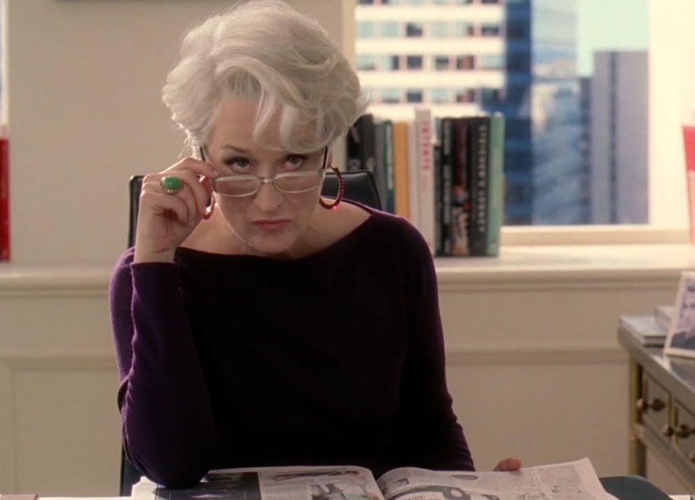 A woman with short white hair, wearing glasses and a dark top, sits at a desk holding a magazine and looks up over her glasses with a serious expression. Books and a window with city buildings are in the background.