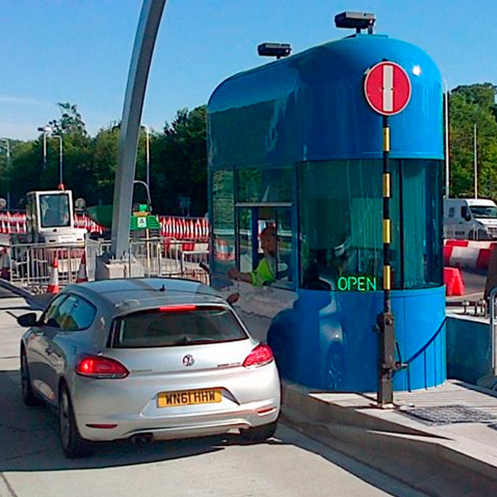 A silver car waits at a blue toll booth displaying a green "OPEN" sign. A worker is visible inside the booth, and there are traffic barriers and roadwork in the background.
