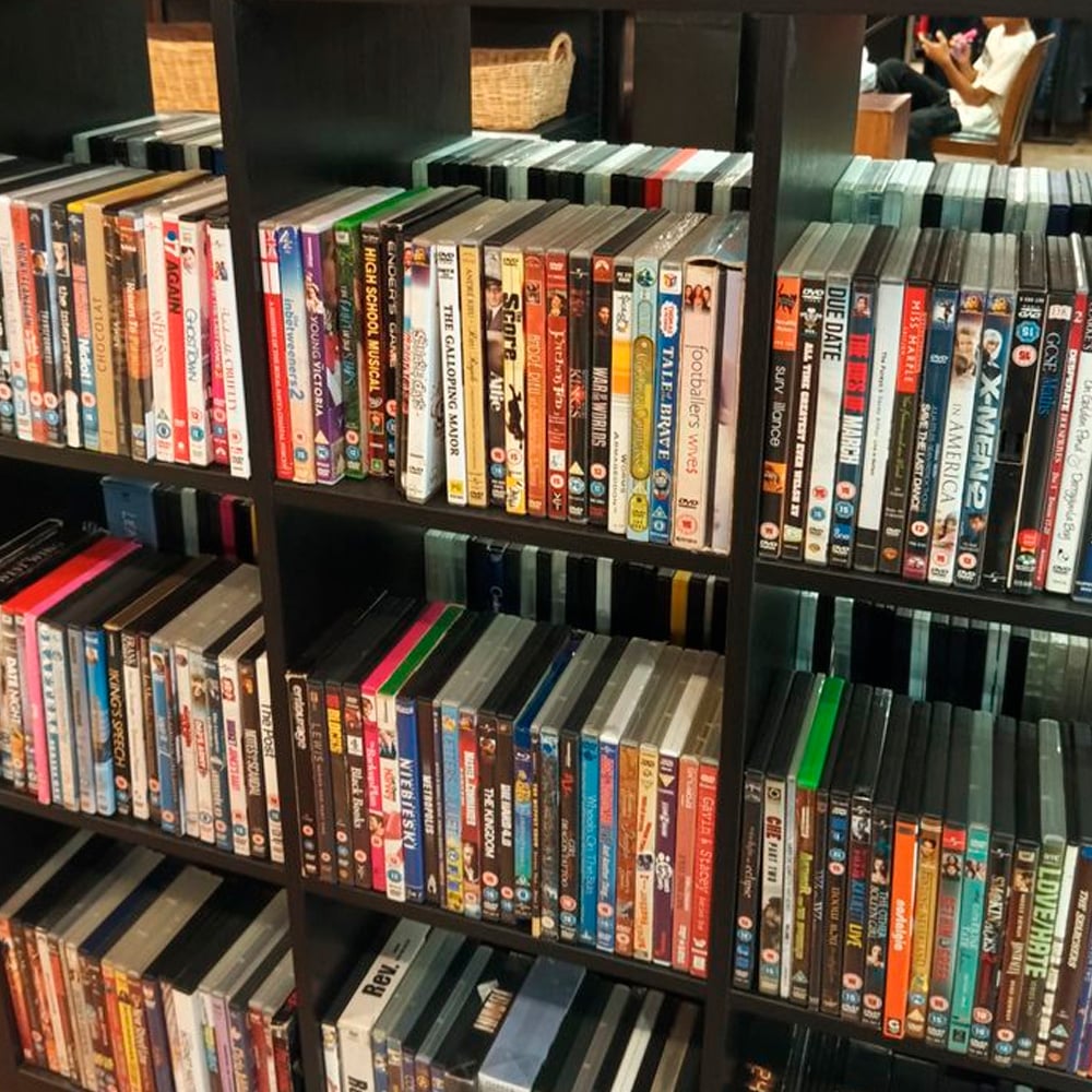 Shelves filled with rows of assorted DVDs in colorful cases, neatly organized side by side. Some baskets and a reflection of people sitting are visible in the background.