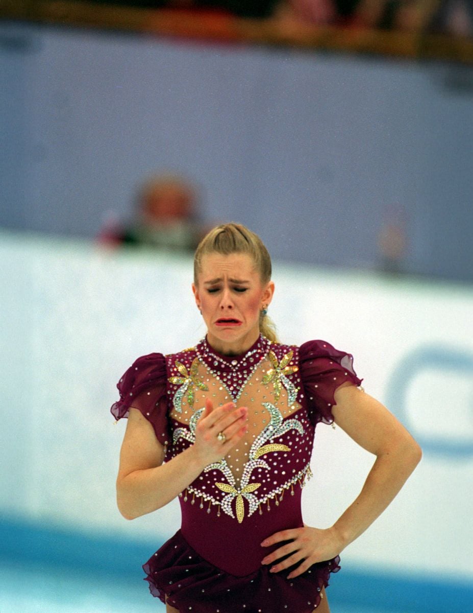 A figure skater in a maroon costume stands on the ice, looking distressed with a tearful expression, holding one hand near her chest during a competition.