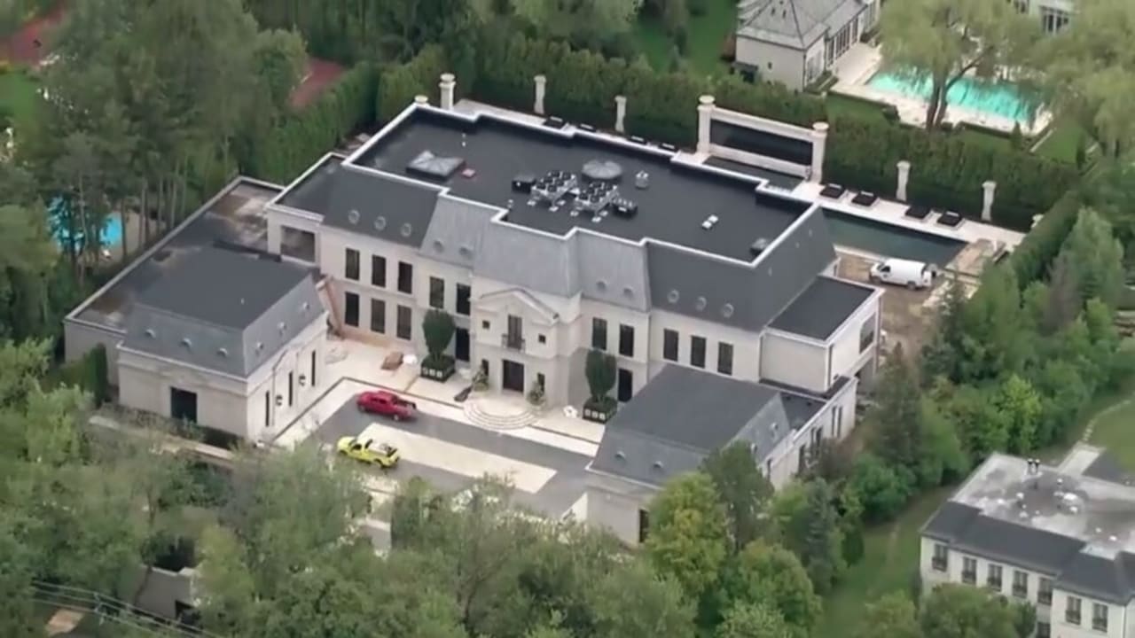 Aerial view of a large, elegant mansion with a black roof, spacious driveway, and two parked cars, surrounded by trees and neighboring houses with pools.