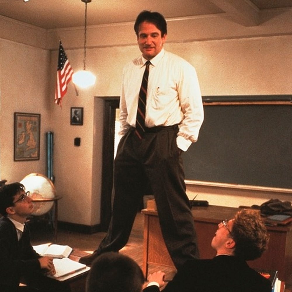 A man in a shirt and tie stands on a desk in a classroom, addressing seated students who look up at him. An American flag, chalkboard, and globe are visible in the background.
