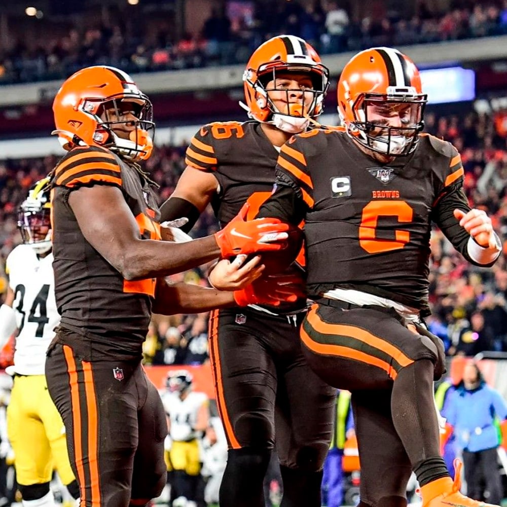 Three Cleveland Browns football players in brown and orange uniforms celebrate a touchdown on the field, with one player holding the ball and fans visible in the stadium background.