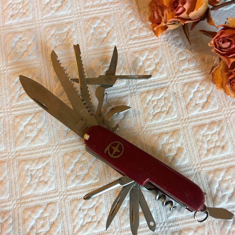 A red Swiss Army knife with multiple tools extended lies on a white, patterned tablecloth near a bunch of dried orange roses in the top right corner.