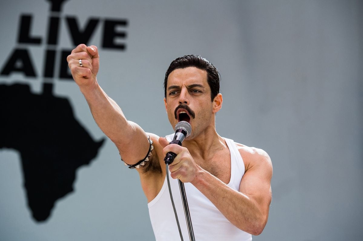 A man in a white tank top holds a microphone and raises his fist while performing on stage, with the word "LIVE" and part of an Africa map visible in the background.