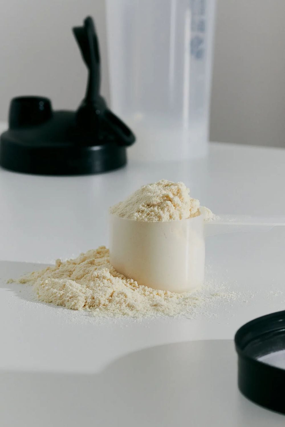A white plastic scoop filled with protein powder sits on a white surface, with some powder spilled around it. A black shaker bottle lid and a clear shaker bottle are in the background.