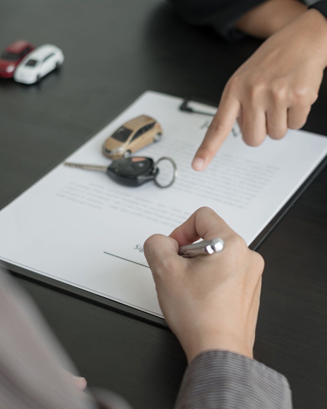 A person is about to sign a car sales contract on a clipboard, while another hand points to the signature line. Car keys and small toy cars are on the table.