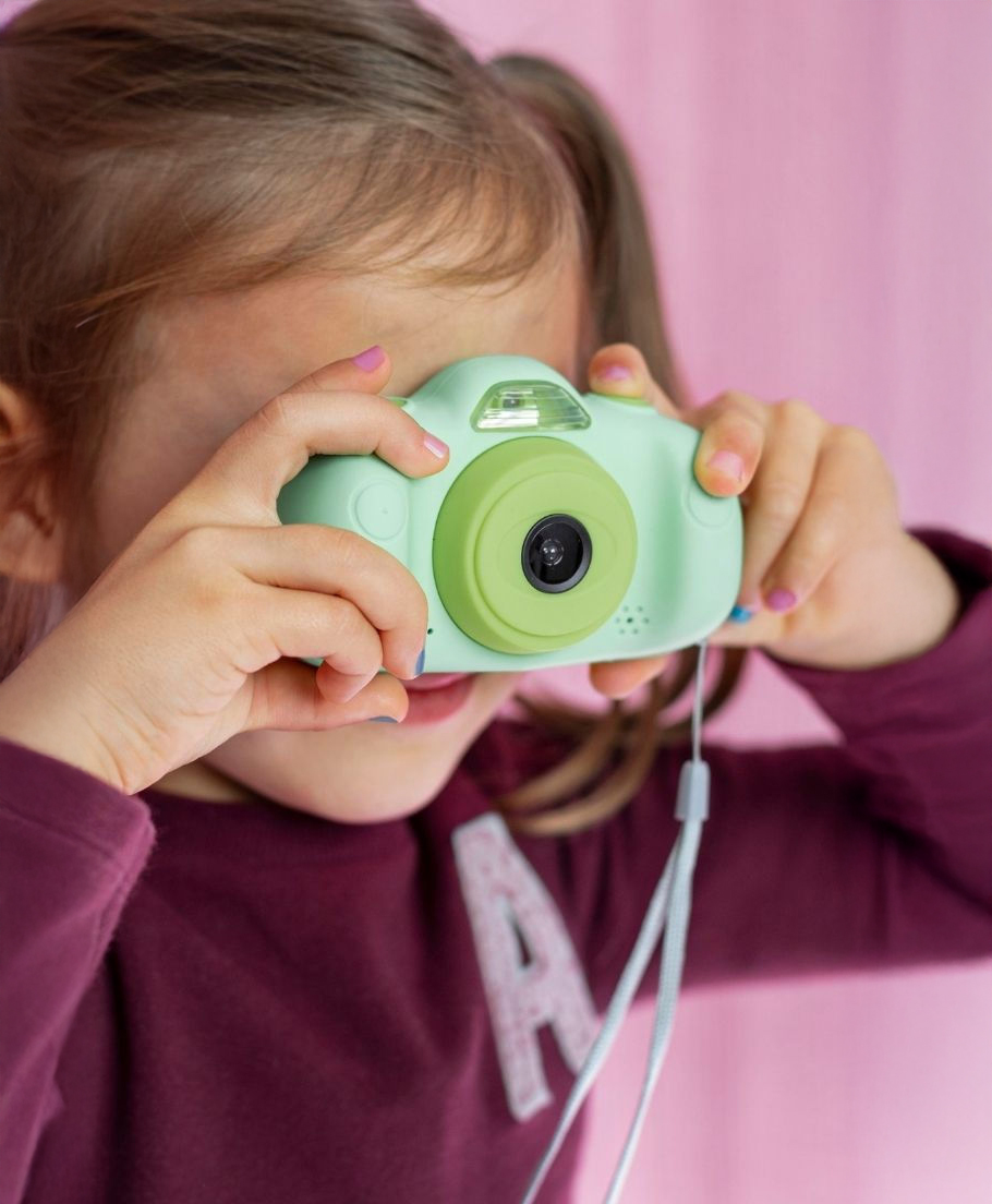 A young child in a purple shirt holds a green toy camera up to their face, pretending to take a photo in front of a pink background.