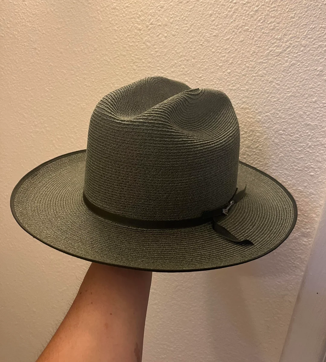 A hand holds a wide-brimmed, textured gray hat with a black band and bow, in front of a cream-colored textured wall.