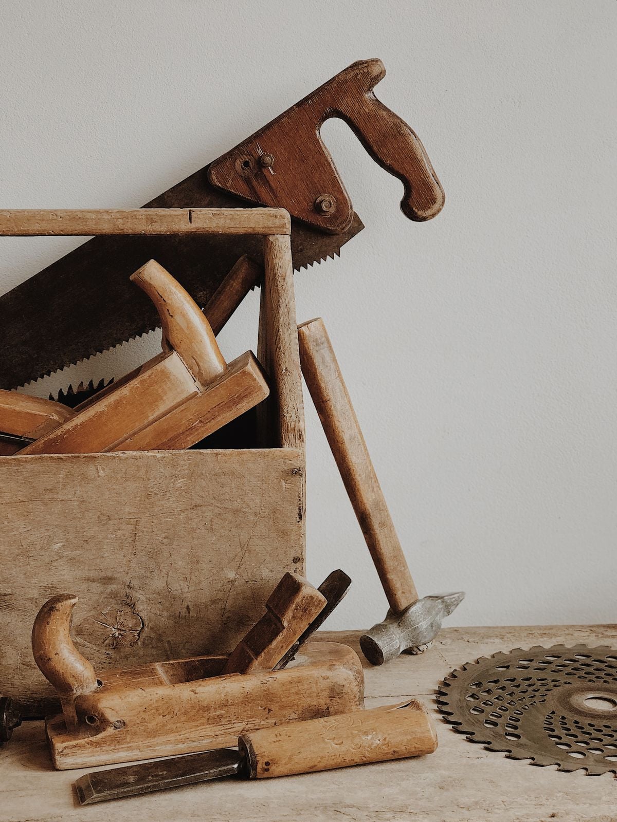 A wooden toolbox filled with vintage hand tools, including a saw, wooden planes, and a hammer, sits on a wooden surface next to a circular saw blade, all against a plain white wall.