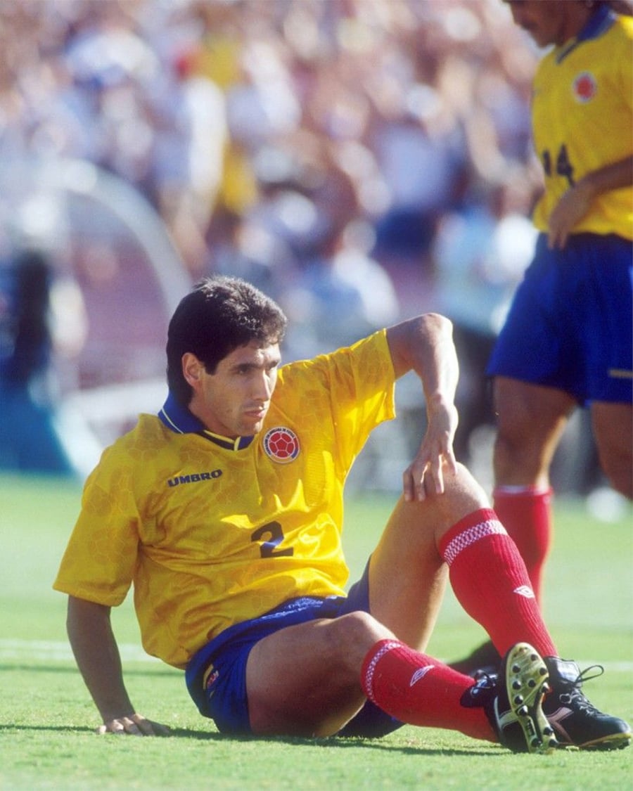 A soccer player in a yellow Colombia jersey and red socks sits on the grass field, looking to the side, during a match. Another player stands in the background.