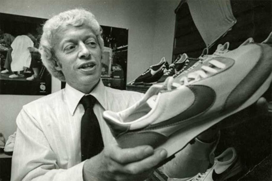 A man with curly hair and a tie holds up a sneaker in a room filled with athletic shoes, examining it closely. Posters of sneakers are visible on the wall behind him.
