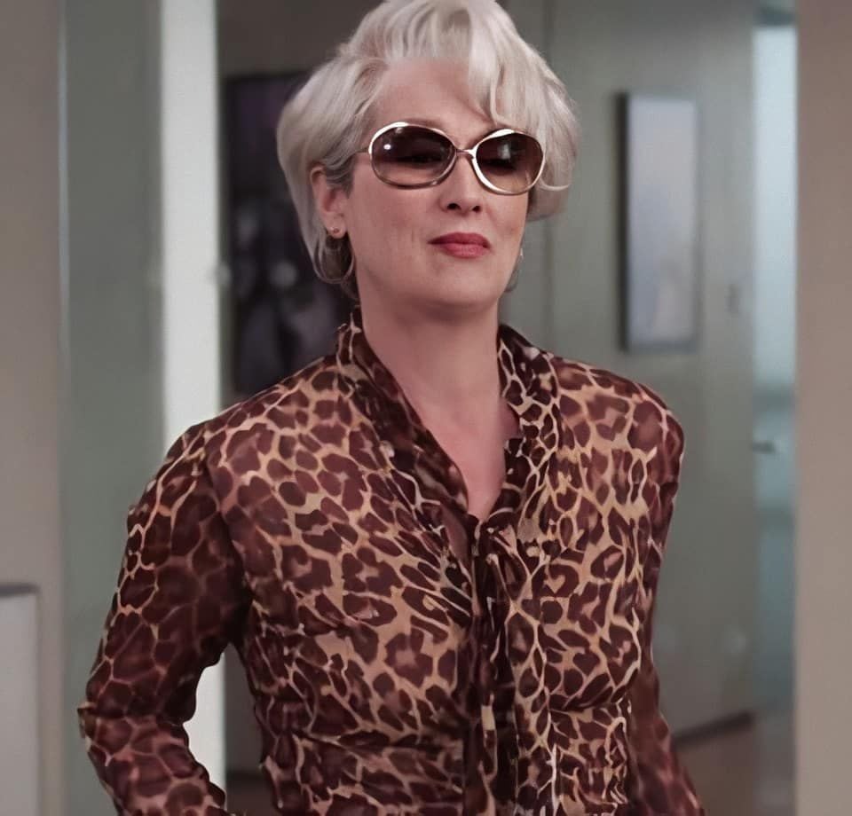A woman with short, silver hair wearing sunglasses and a leopard print blouse walks confidently in an indoor setting.