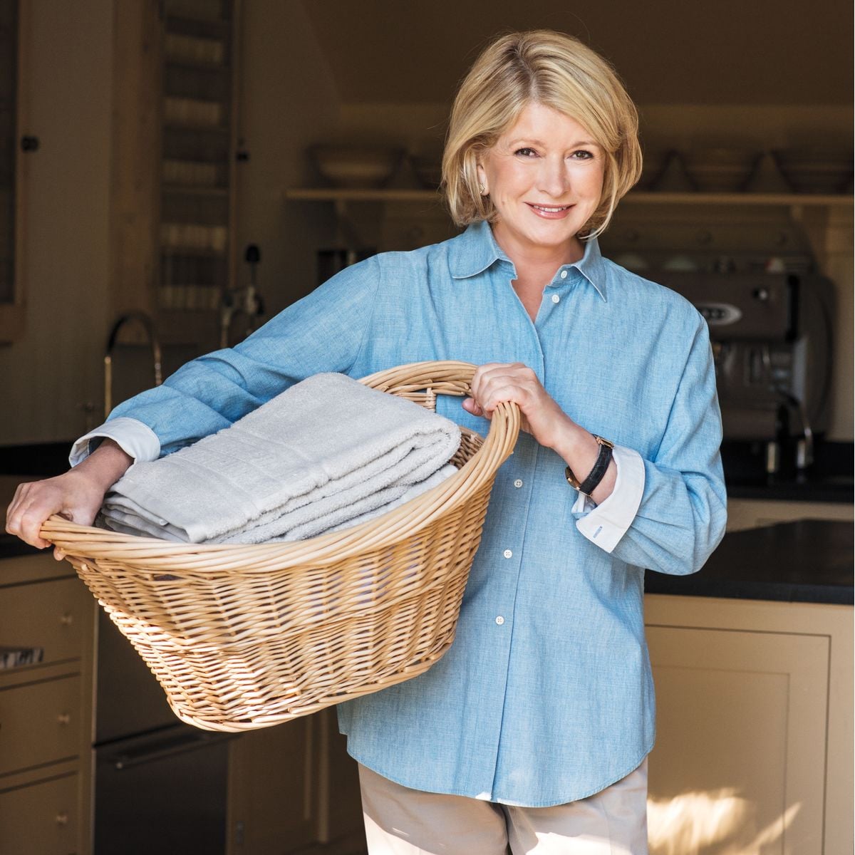 A woman wearing a light blue shirt smiles while holding a wicker laundry basket filled with neatly folded towels in a kitchen or laundry room setting.