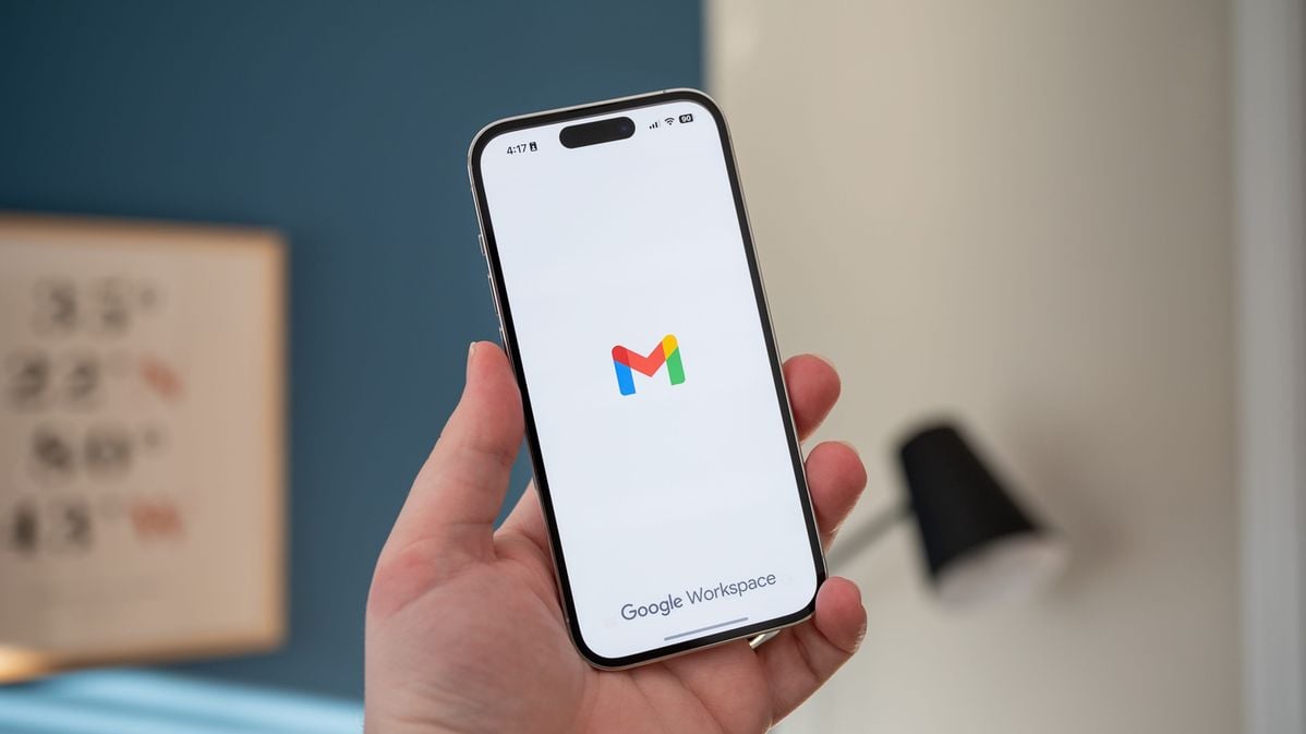 A hand holds a smartphone displaying the Gmail app logo on the screen, with "Google Workspace" text at the bottom. The background is indoors with blurred decor.