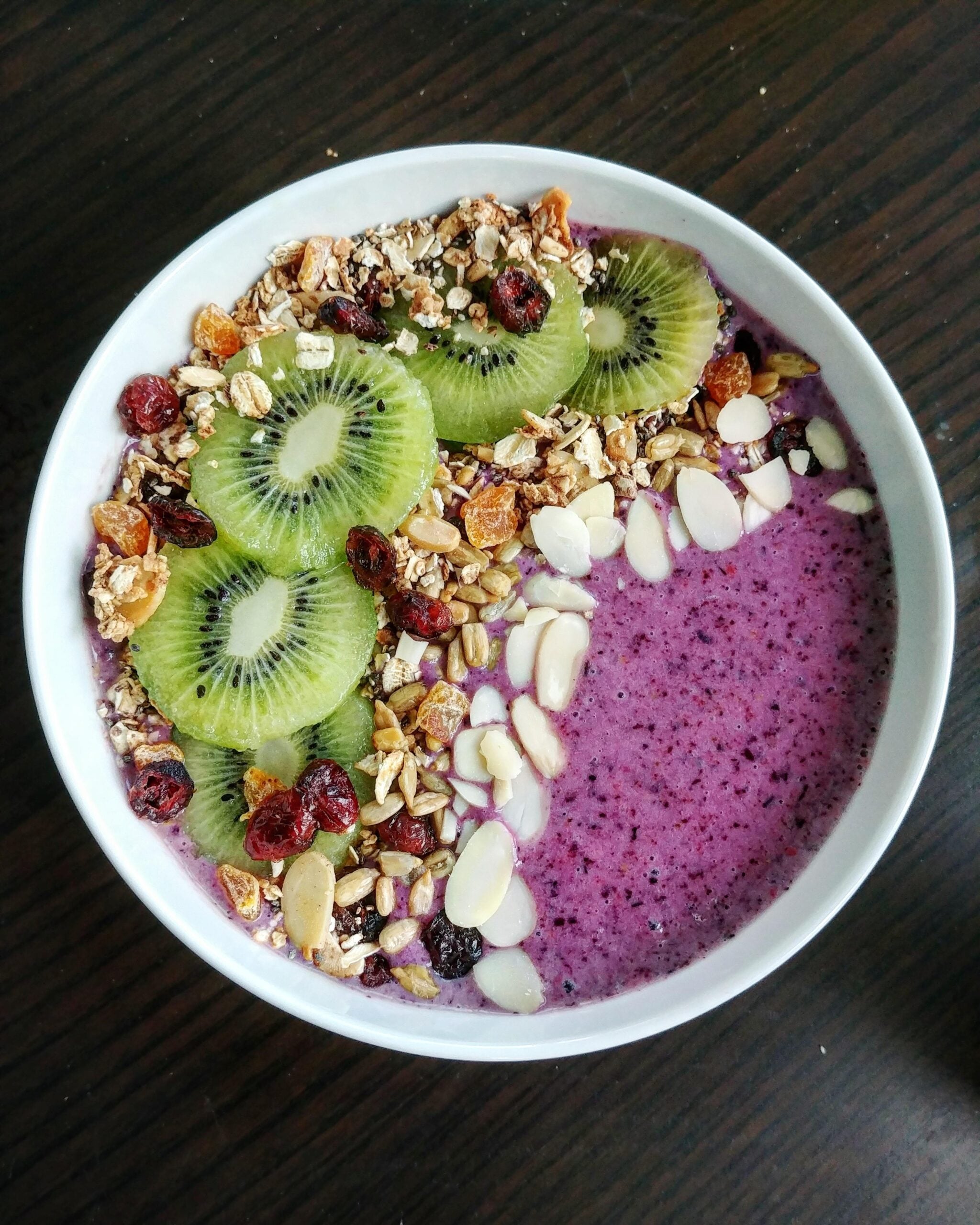 A smoothie bowl with purple blended base, topped with sliced kiwi, granola, dried cranberries, and almond slivers, served in a white bowl on a dark wooden table.