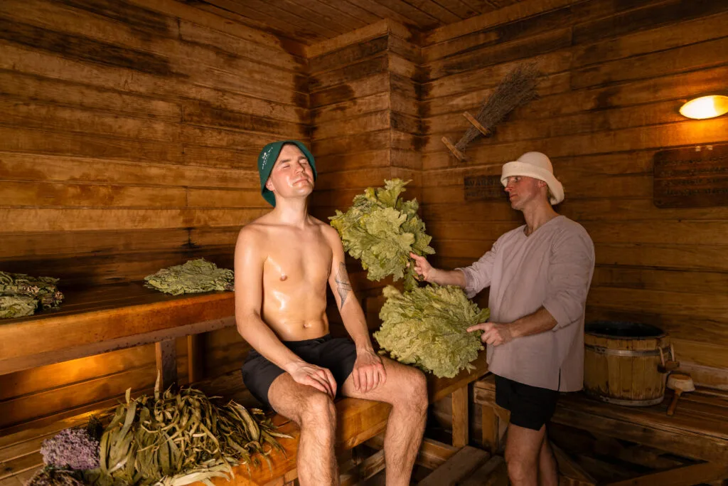 Two men in a wooden sauna; one sits shirtless on a bench with eyes closed, wearing a hat, while the other stands and gently hits him with birch branches in a traditional sauna ritual.