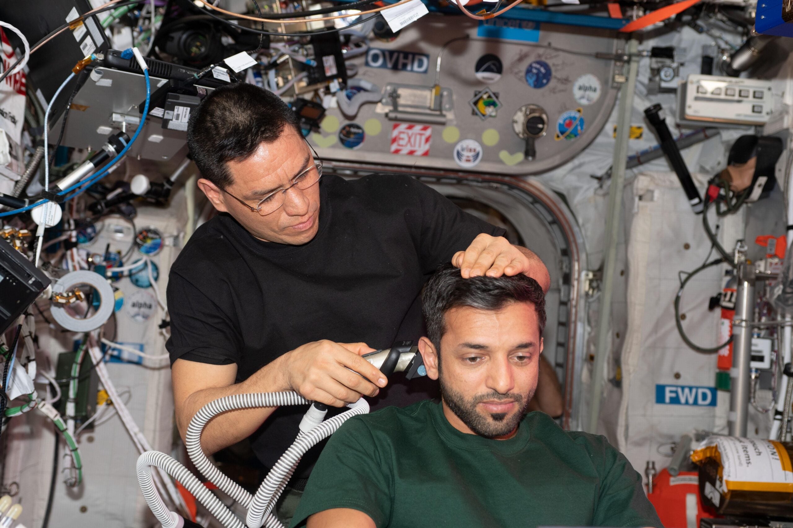 An astronaut gives another astronaut a haircut using electric clippers inside a space station surrounded by various equipment and control panels.