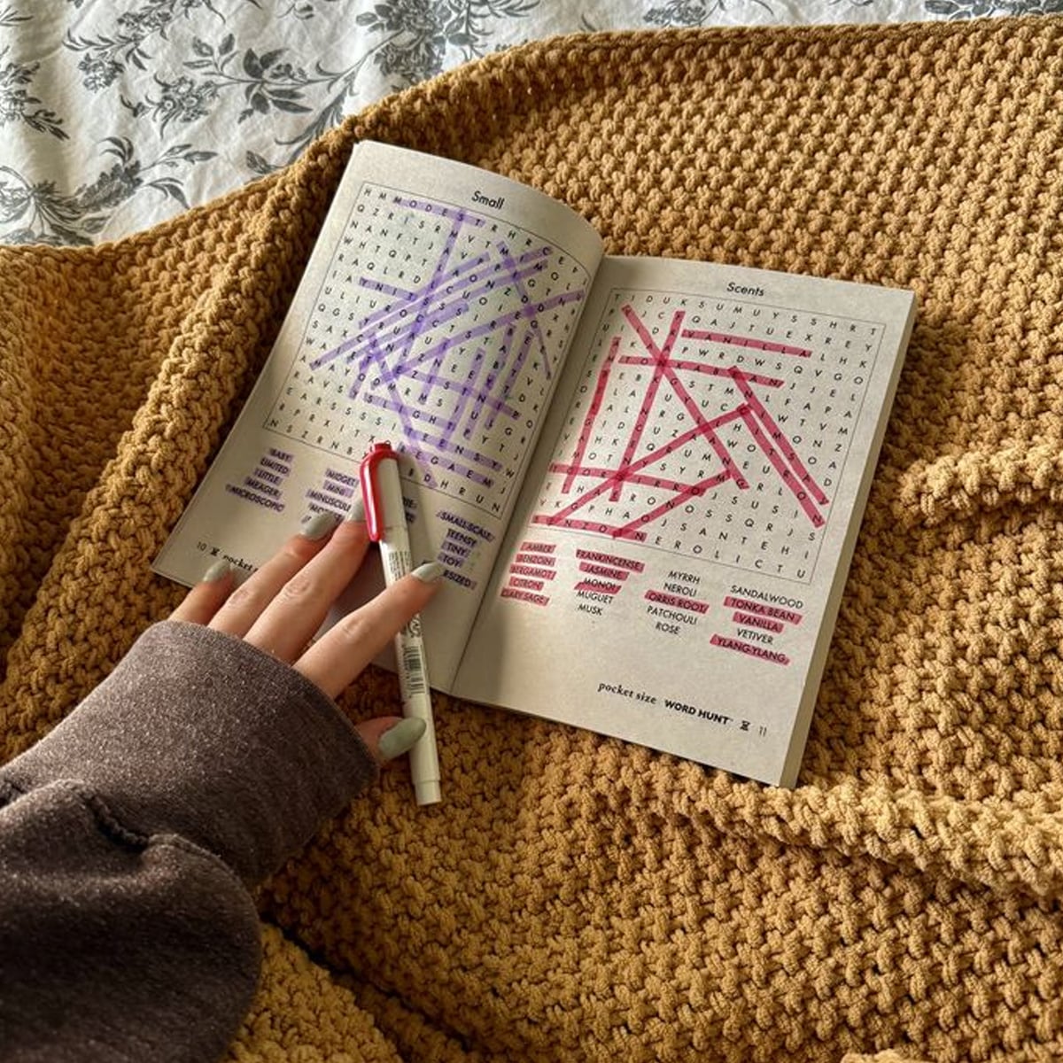 A hand holding a red pen rests on an open word search puzzle book placed on a chunky yellow knit blanket. Several words are highlighted and crossed out in purple and pink.