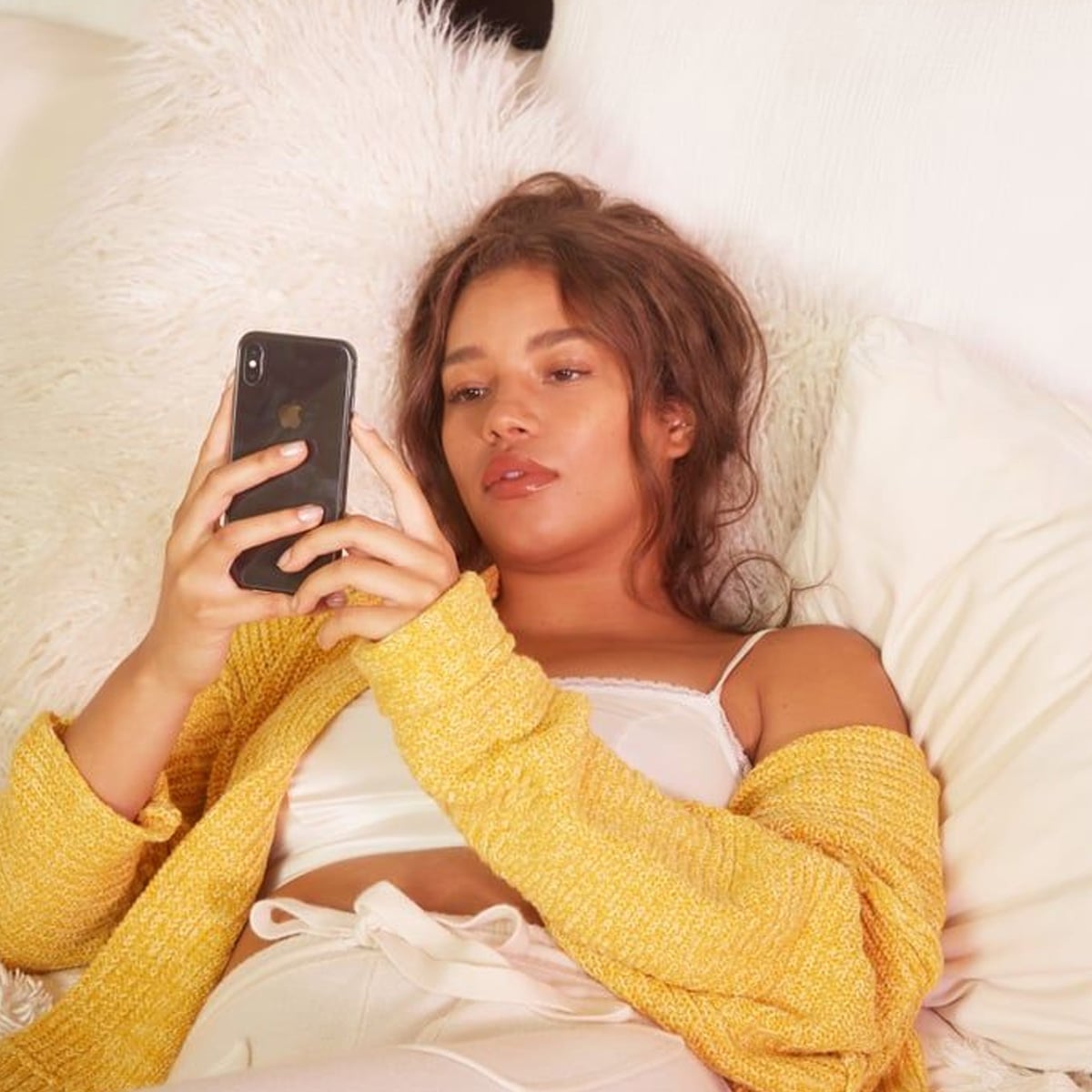 A woman lies on a bed with white pillows and a fluffy blanket, wearing a yellow cardigan and white clothing, looking at her smartphone with a relaxed expression.