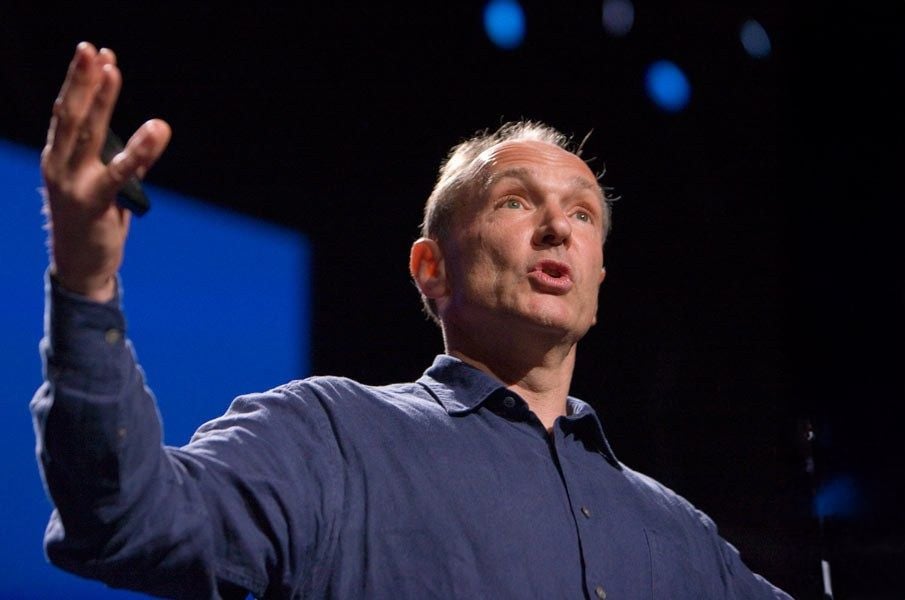 A man in a blue shirt gestures with one hand raised while speaking on stage, with a dark background and blue lights behind him.