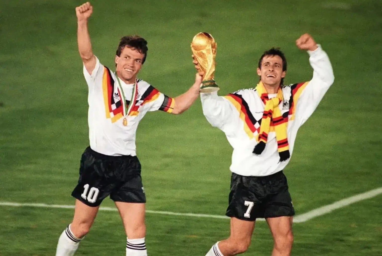 Two soccer players in white jerseys with black, red, and yellow stripes celebrate on a field, one holding up a gold trophy. Both have medals around their necks and are raising their arms in victory.