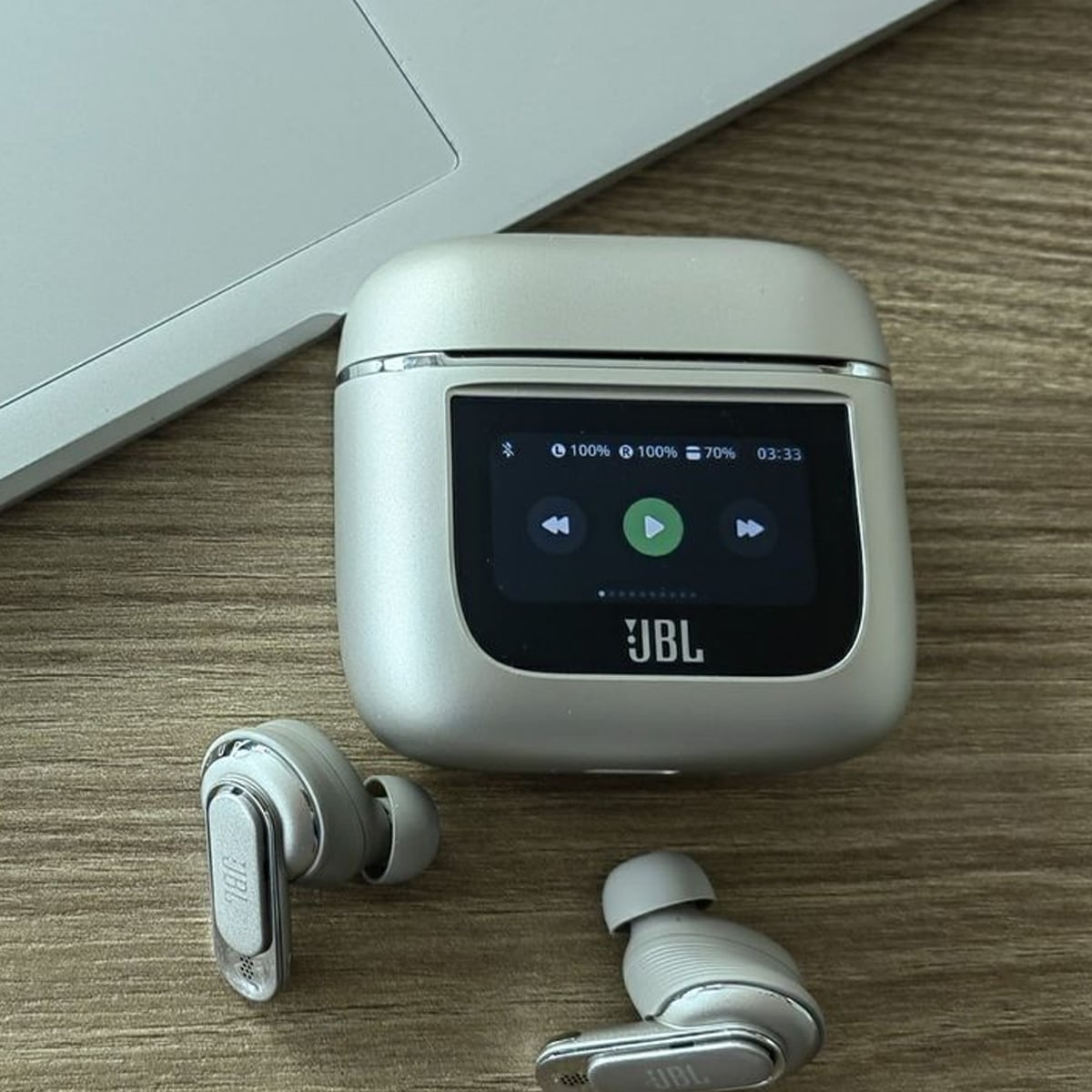 A pair of gray JBL wireless earbuds rests beside their charging case, which features a display screen showing playback controls and battery levels. Part of a silver laptop is visible on the wooden surface.