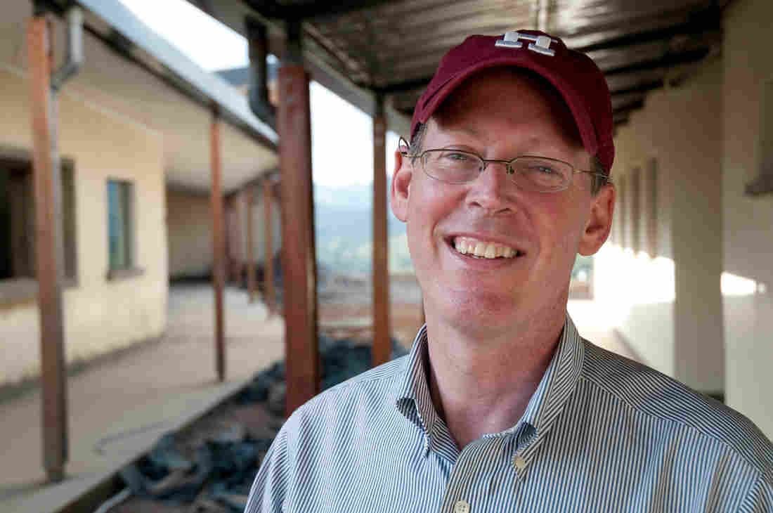 A man wearing glasses, a maroon baseball cap, and a striped shirt smiles at the camera while standing in a covered outdoor walkway with buildings in the background.