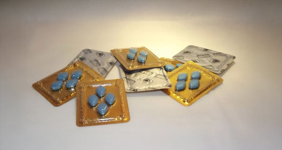 Blister packs containing blue, diamond-shaped pills are scattered on a light surface. Some packs are partially open, revealing the pills, while others remain sealed in silver and gold packaging.