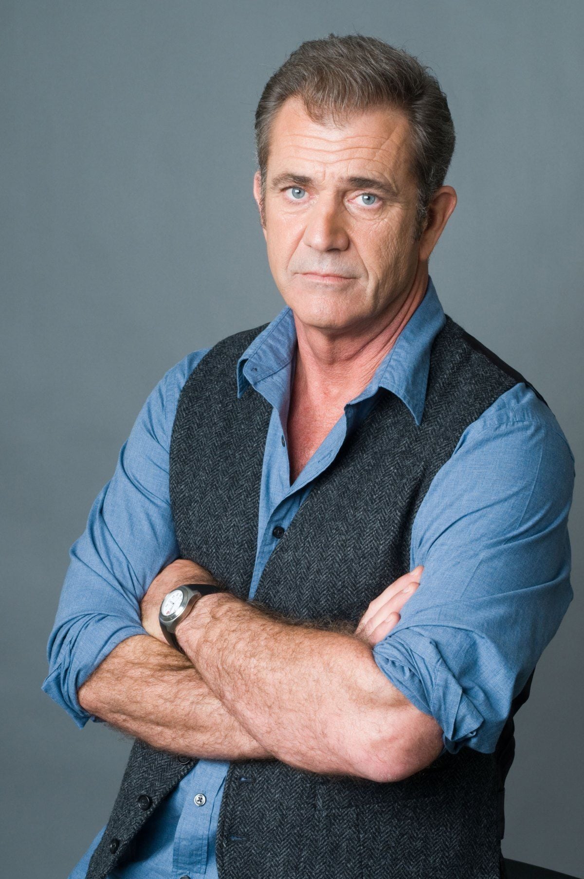 A middle-aged man with short, grayish hair wears a blue button-up shirt and a dark vest. He stands with arms crossed against a plain gray background, looking directly at the camera with a neutral expression.