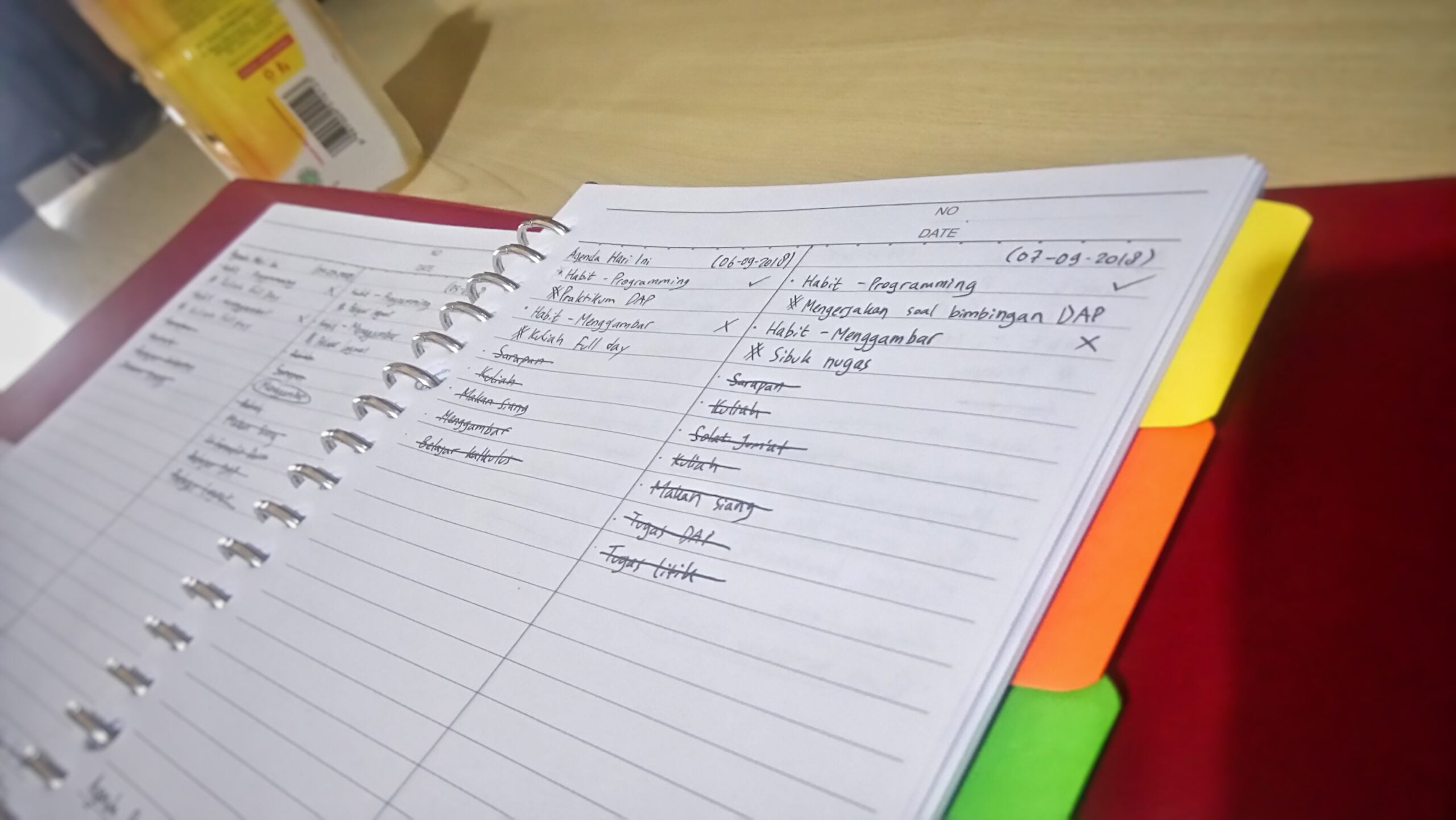A spiral notebook with handwritten notes and checklists lies open on a desk, with colorful tab dividers visible on the right. A beverage carton is partially seen in the upper left corner.
