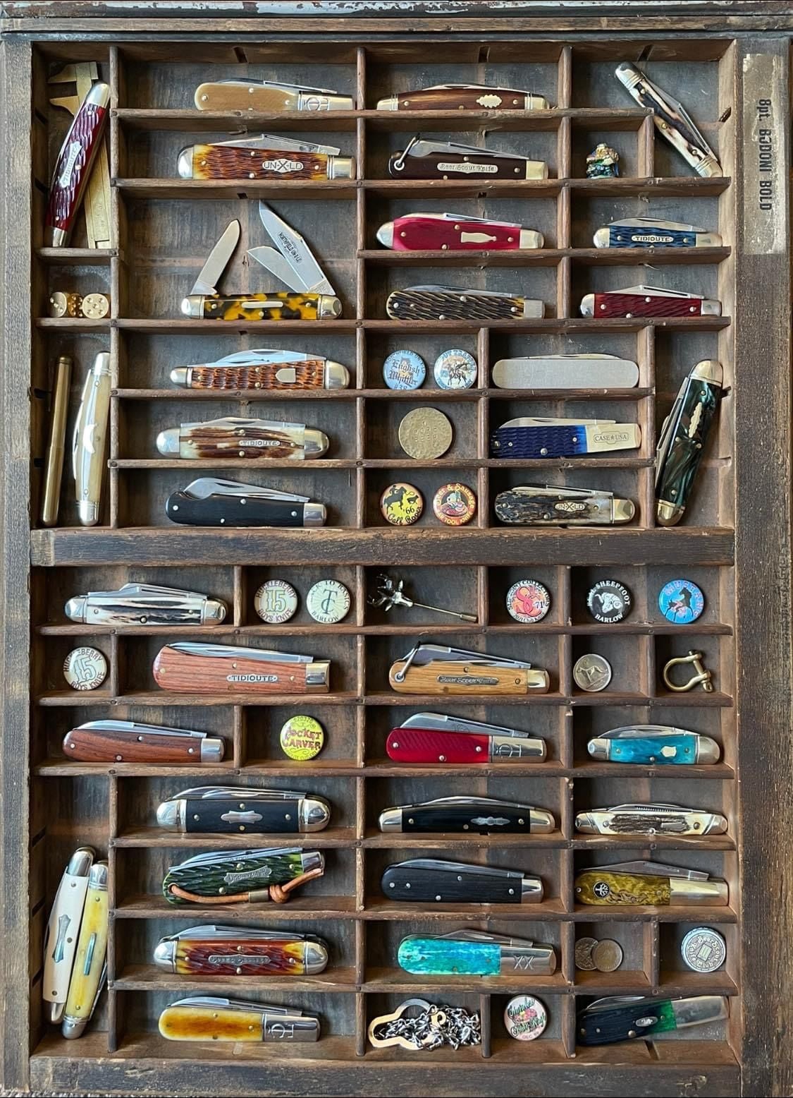 A wooden display case holds a neatly arranged collection of pocket knives, vintage pins, coins, a key, a whistle, and a pocket watch, each item separated in its own compartment.