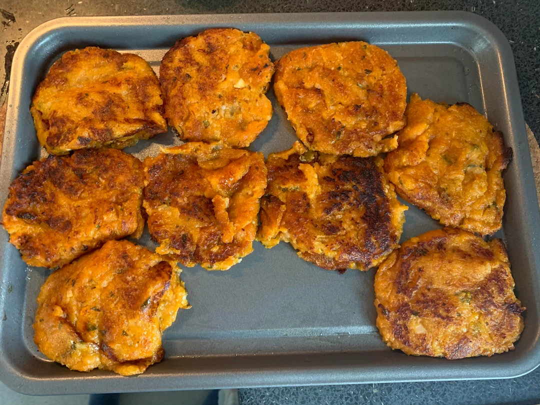 A baking tray with nine golden-brown, irregularly shaped sweet potato patties, slightly crispy on the outside, resting on a dark countertop.