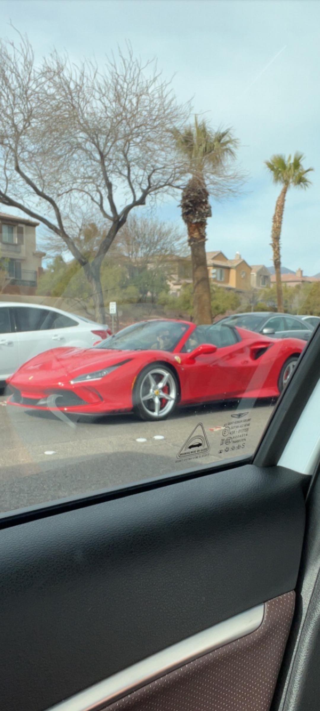 A red convertible sports car is parked on the street near palm trees and other cars, viewed through the window of a nearby vehicle. Houses and a clear sky are visible in the background.