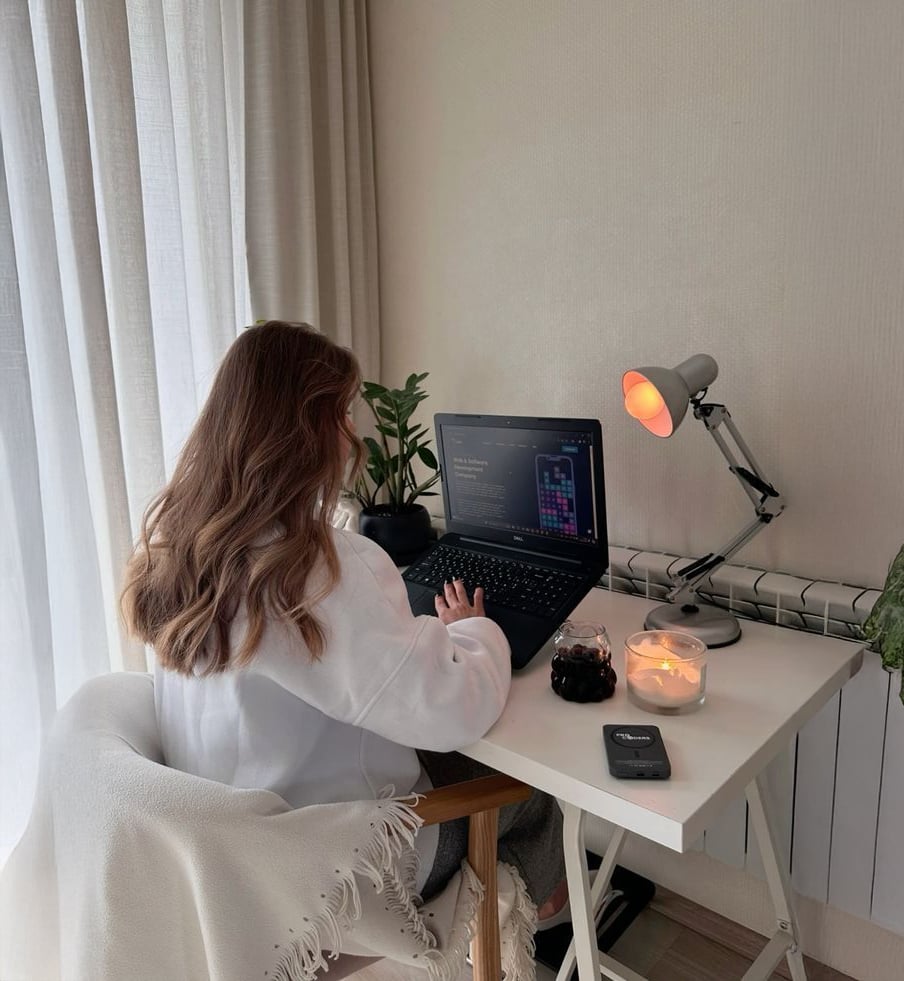 A person with long hair sits at a desk working on a laptop in a cozy room. The desk has a lit candle, a smartphone, a plant, a lamp, and a glass of a dark beverage. Light filters through sheer curtains nearby.